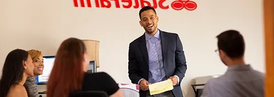 A male speaker, standing in front of a State Farm logo, addresses his fellow workers.